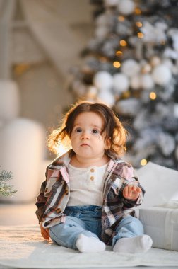 Beyaz halıda oturan sevimli bir kız bebek. Ekose gömlek giyiyor. Noel kutlamaları boyunca kapalı bir yerde, parlak ışıklarla.