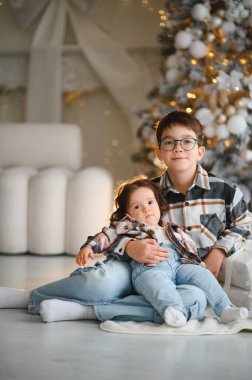 Erkek ve kız kardeş yerde birlikte oturup Noel tatilinin tadını çıkarıyorlar. Neşeli ışıklarla aile portresi