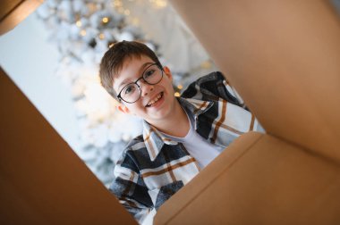 Gözlüklü çocuk gülümsüyor, kahverengi bir kutuya bakıyor, bayram döneminde bir hediyeyi açıyor.