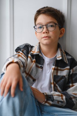 Kendine güveni olan mavi gözlü, modaya uygun çerçeveler takan, günlük giysiler giyen, açık renkli bir duvara yaslanmış bir çocuk.