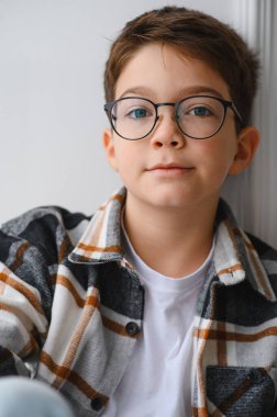 Modern gözlüklü ve ekose gömlekli bir çocuk kameraya dostça bir ifadeyle bakıyor.
