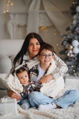 Bir battaniyeye sarılı gülümseyen bir anne, oğul ve bebek ile aile portresi, Noel tatilinde samimi bir bağ kurma anı paylaşıyor.