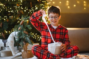 Gülümseyen genç çocuk, Noel ağacının yanında bir bayram hediyesi açıyor, sevinç ve heyecanı ifade ediyor.