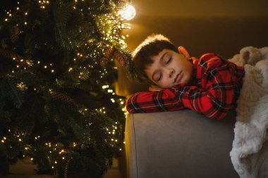 Tatil hayali kuran çocuk, ışıklarla aydınlatılmış bir Noel ağacının yanında huzur içinde dinleniyor.