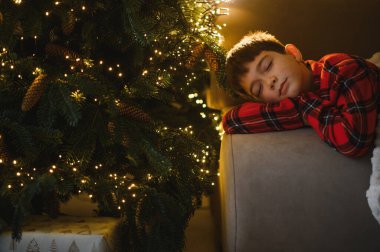 Genç çocuk bir kanepede sessizce dinleniyor, bayram hediyelerini düşlerken Noel ağacı süsleniyor.