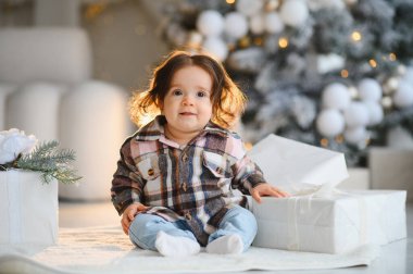 Mutlu bebek beyaz noel hediyeleriyle halının üzerinde oturuyor ve arka planda bulanık bir festival ağacı, tatil sezonunu kutluyor.