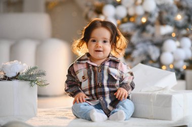 Ekose gömlek ve kot pantolon giyen tatlı çocuk Noel hediyeleriyle beyaz bir halıya oturmuş, bayram sezonunu kutluyor.
