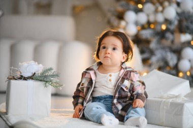 Bebek bebek halının üzerinde beyaz noel hediyeleriyle oturuyor, evin içinde süslü bir ağacın yanında şaşkınlıkla yukarı bakıyor.