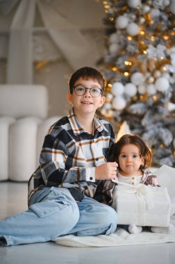 Genç erkek ve kız kardeş, kış tatilinde birlikte bir hediye açıyor, süslü bir ağacın yanında yerde oturuyor.