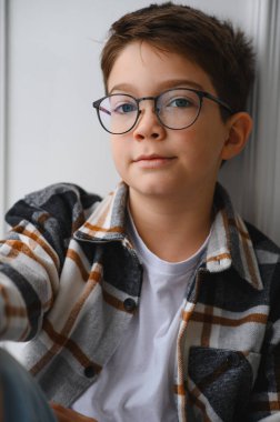 Yuvarlak gözlüklü ve ekose gömlekli genç bir öğrenci, özenle beyaz arka planlı bir stüdyo fotoğrafıyla kameraya bakıyor.