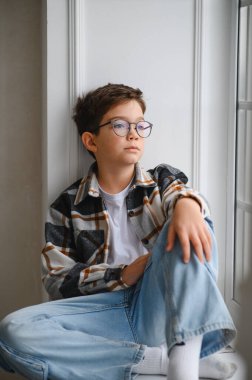 Gözlüklü ve ekose gömlekli genç bir çocuk, pencerenin kenarında oturuyor ve düşünceli bir şekilde, dalgın dalgın dalgın ya da hayal kuruyor.