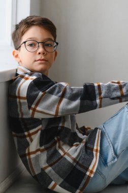 Ekose gömlekli ve kotlu bir çocuk, duvara yaslanmış, doğrudan izleyiciye bakıyor. Düşünceli ve sıradan çocukluk anı.
