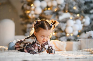 Evdeki yumuşak beyaz halıda gülümseyen ve oynayan küçük bir kız, Noel ağacının yanında sıcak bir kış tatili geçiriyor.
