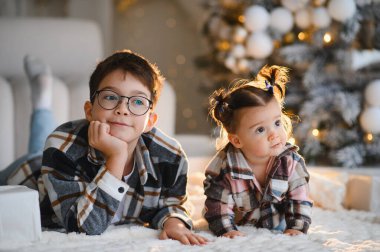Kardeşler yumuşak bir halıya uzanmış, Noel ağacı ışıklarına hayretle bakıyor, birlikte mutlu anları kutluyorlar.