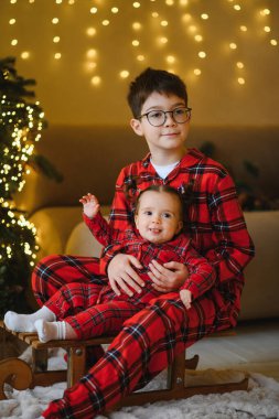 Kardeşini elinde tutan, aynı kırmızı pijamaları giyen, Noel tatili boyunca tahta bir kızağın üzerinde oturan kardeş.