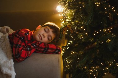 Genç bir çocuk rahat bir şekilde kanepede dinleniyor, tatil gecelerinde rüya görüyor, süslenmiş bir Noel ağacından gelen hafif şenlik ışıklarıyla aydınlanıyor.