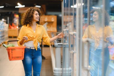 Kıvırcık saçlı bir kadın markette dondurucunun kapısını çekerek açıyor. Elinde turuncu bir alışveriş sepeti var.