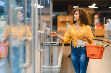Bir markette dondurucudan dondurulmuş ürünler seçen bir kadın elinde sebzeli bir alışveriş sepeti tutuyor.