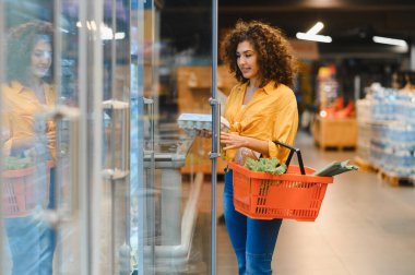 Alışveriş sepetli bir kadın markette buzdolabından yumurta seçiyor, taze gıda ürünleri alıyor.