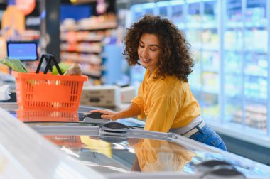 Kıvırcık saçlı genç bir kadın markette dondurucudan bir şeyler topluyor. Yakınlarda turuncu bir alışveriş sepeti var.