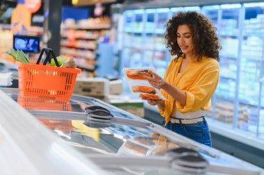 Bir marketteki dondurucudan paketlenmiş somon seçen kadın müşteri sağlıklı beslenmek için taze deniz ürünleri alıyor.