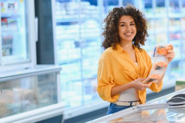 Gülümseyen ve paketlenmiş somon filetoları tutan bir kadın, marketteki dondurucu koridorundan sağlıklı deniz ürünlerini seçen bir kadın.
