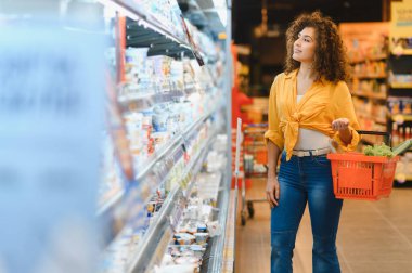 Bir kadın, modern bir perakende mağazasından alışveriş yaparken elinde bir alışveriş sepeti tutarak soğutulmuş raftan ürünler seçiyor.