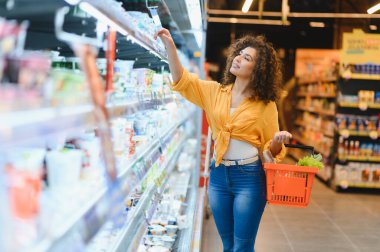 Elinde alışveriş sepeti olan bir kadın, bir markette parlak ışıklandırılmış dondurulmuş bir vitrinden ürünleri seçiyor ve araştırıyor.