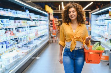 Bakkalda yürüyen kıvırcık saçlı genç bir kadın içi taze ürünlerle dolu turuncu bir alışveriş sepeti taşıyor.