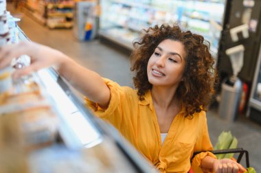 Genç bayan müşteri raftaki ürünlere uzanıyor, alışveriş merkezindeki sepeti için eşyalar seçiyor.