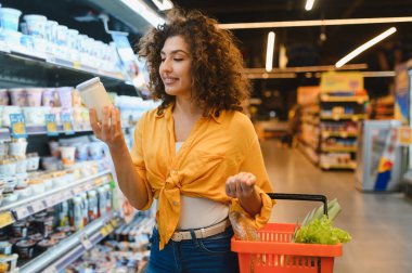 Bakkal alışverişi yapan genç bir kadın, mandıra koridorundaki yoğurt etiketini dikkatle okuyarak sağlıklı yiyecek seçimlerine öncelik veriyor.