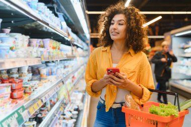 Bir kadın süpermarket koridorunda duruyor, telefonunu ve alışveriş sepetini tutuyor, süt ürünlerini karıştırıyor.