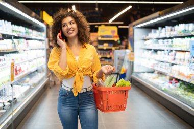 Portakal sepeti içinde gülümseyen, sebze alışverişi yapan ve markette cep telefonuyla konuşan genç bir kadın.