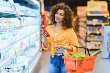 Elinde akıllı telefon ve sepet olan bir kadın, markette alışveriş yaparken alışveriş listesiyle eşyaları karşılaştırıyor.
