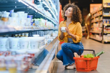 Çömelmiş bir kadın, elinde yoğurt kabıyla marketin soğutulmuş süt ürünleri reyonundan alışveriş yapıyor.