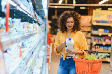 Elinde süt şişesi olan bir kadın marketteki soğutmalı koridordan elinde bir sepetle süt ürünleri seçiyor.