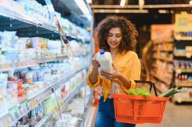 Elinde bir şişe süt tutan bir kadın markette elinde sepetle market alışverişi yaparken ürün etiketini kontrol ediyor.
