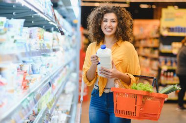 Mutlu kadın yiyecek alışverişi yapıyor, elinde bir şişe süt ve bir sepet dolusu taze ürünle modern bir markette