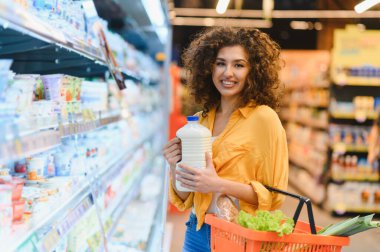 Gülümseyen, elinde süt şişesi ve alışveriş sepeti olan bir kadın süpermarketten alışveriş yapıyor. Günlük ev işleri ve sağlıklı yaşam tarzı.