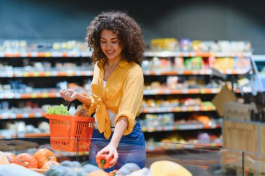 Elinde alışveriş sepeti olan genç bir kadın marketten taze kırmızı dolma biber seçiyor, sağlıklı beslenmeye odaklanıyor.