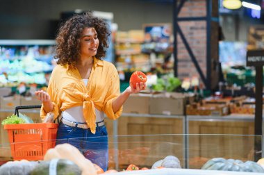 Gülümseyen genç bir kadın sebze reyonundan taze sebze seçiyor, marketten sağlıklı yiyecek alıyor.