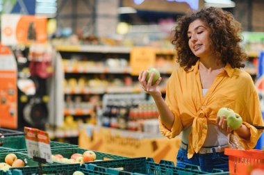 Genç kıvırcık saçlı kadın dikkatle meyve topluyor, yoğun bir mağaza koridorunda sağlıklı beslenme ve market alışverişine odaklanıyor.
