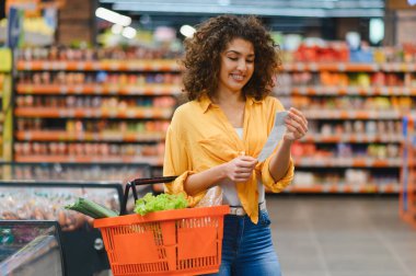 Market raflarının önünde elinde bir sepet taze yiyecekle uzun bir alışveriş makbuzunu incelerken gülümseyen genç bir kadın.