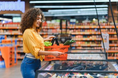 Dondurucudan bukleli saç toplayan genç bir kadın markette elinde taze sebzelerle bir alışveriş sepeti tutarken