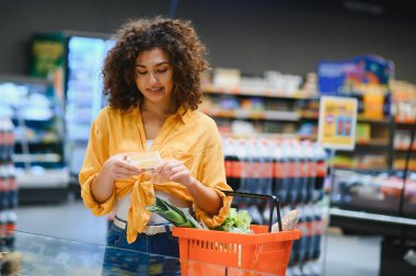 Bir markette alışveriş yaparken elinde turuncu bir sepetle peynir paketini dikkatlice inceleyen bir kadın.