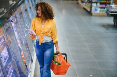 Bir markette dondurucudan ürün seçerken elinde bir paket yiyecek ve turuncu alışveriş sepeti tutan bir kadın.