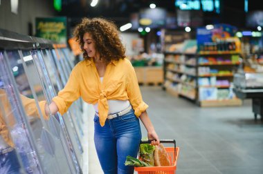 Genç bir kadın süpermarketten alışveriş yapıyor, soğutulmuş bölümden taze şeyler seçiyor ve bir sepet taşıyor.