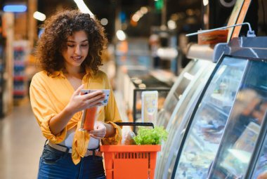 Akıllı telefondan bilgi okuyan bir kadın paketli bir somon balığı ve market alışverişi sepetiyle modern bir süpermarkette