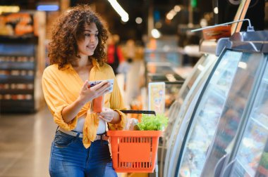 Cep telefonuna bakarken gülümseyen genç bir kadın markette içinde taze sebzeler olan bir alışveriş sepeti tutuyor.