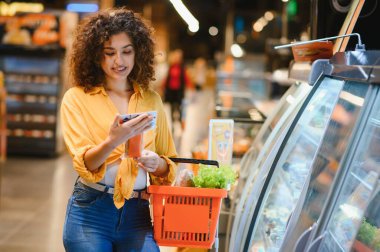 Genç bayan müşteri, elinde turuncu bir alışveriş sepetiyle soğutucudan taze gıda ürünlerini dikkatlice seçiyor.